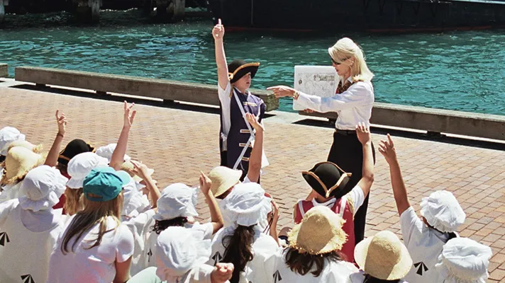 Tour guide in period costume educates children about Sydney Rocks colonial history beside harbour.